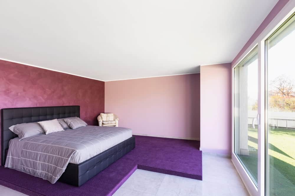 Primary bedroom with a modern and cozy bed set on top of a violet flooring. The room also has a doorway leading straight to the home's backyard.