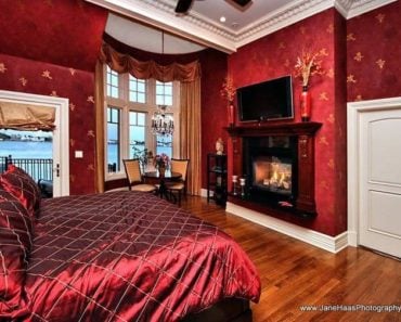 A master bedroom with red decorated walls and an elegant red velvet bed setup with a fireplace and a TV set in front. The room features a sitting area by the windows and a balcony area.