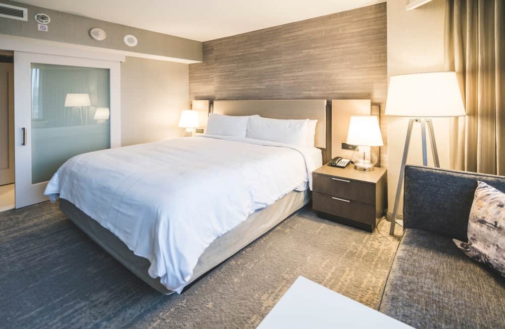 This primary bedroom features a modish bed set lighted by lovely table lamps on bedside tables. The room has stylish flooring and a gray couch on the side.