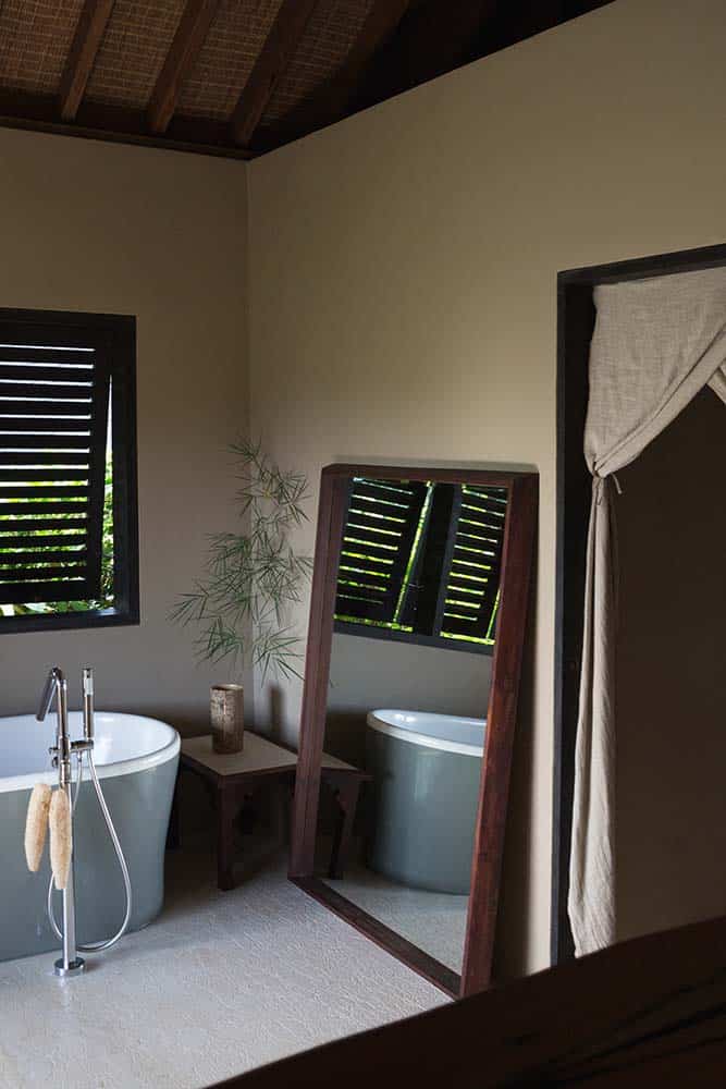 This is a close look at the bathroom with a white porcelain freestanding bathtub across from the wooden vanity sink that matches the frames of the large leaning mirror on the side and the wooden shuttered window. These are then complemented by the potted bamboo plant at the corner.