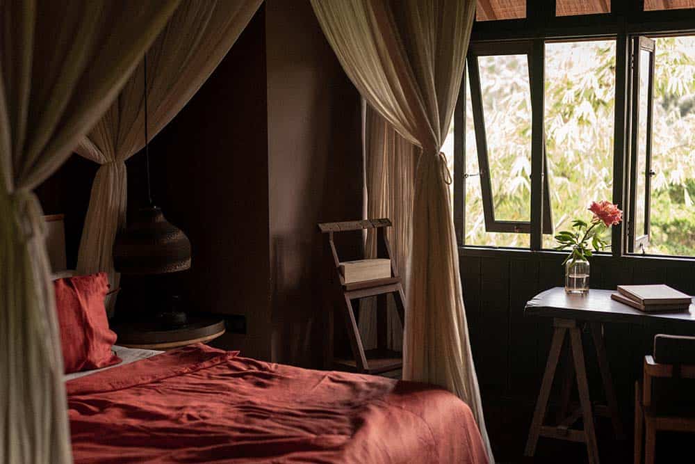 This is the close look of the bedroom with a large bed with salmon sheets surrounded by white curtains hanging from the wooden beamed ceiling adorned by the earthy pendant light on the side and the simple wooden study area by the window.