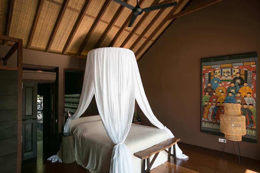 The bedroom has a tall wooden cathedral ceiling that hangs a white curtain around the white bed that stands outa gainst the hardwood flooring. These are then complemented by the colorful artwork on the side and the woven wicker lamp.