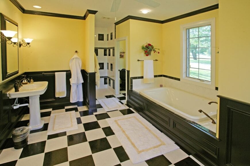 Yellow primary bathroom with black wainscoting and checkered flooring topped by white bordered rugs. It includes a deep soaking tub and a pedestal sink under the framed mirror.