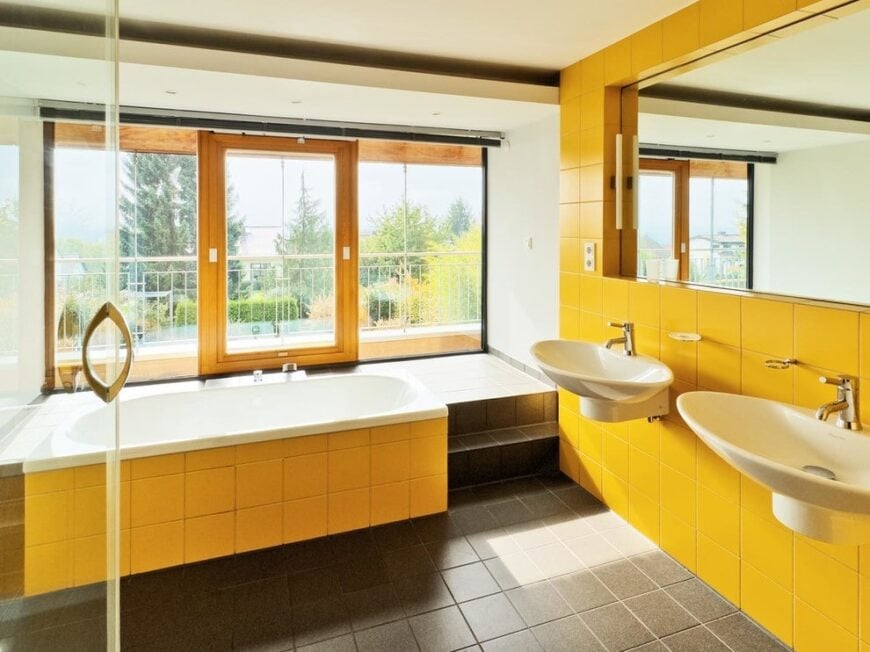 The contemporary primary bathroom boasts a deep soaking tub along with his and her wall mount sinks that are fixed against the yellow tiled wall. It has full height windows and a glazed door leading out to the balcony.