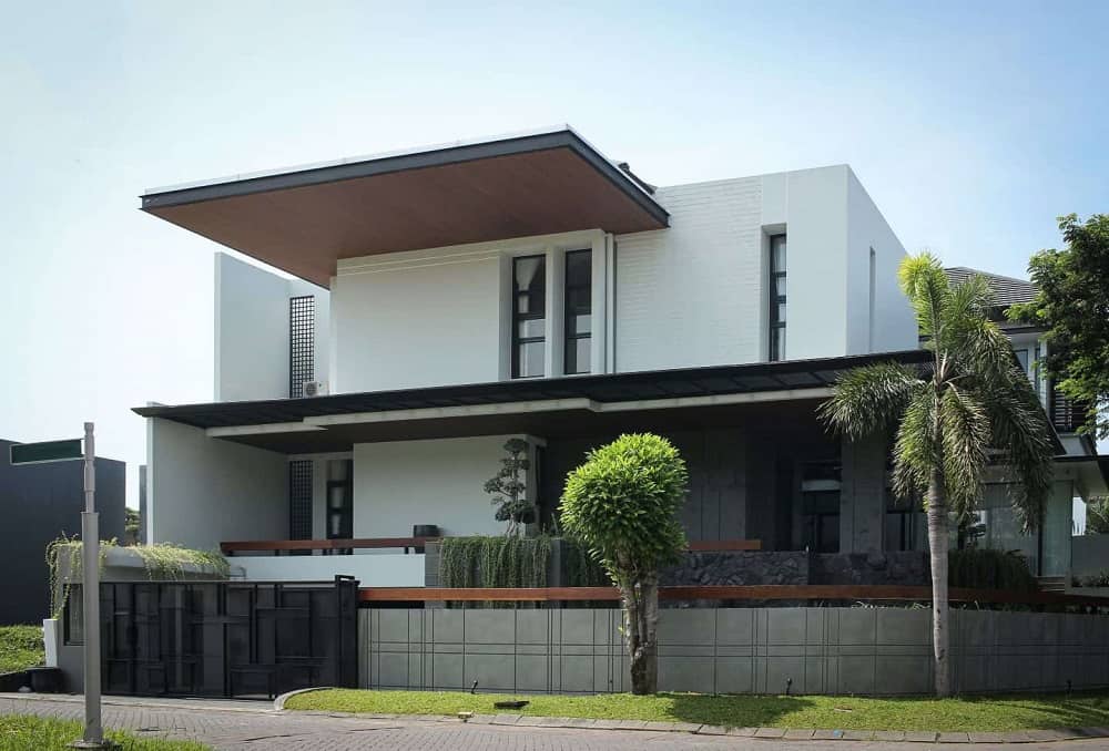 This is a view of the house exterior that has bright white exterior walls, a large roof panel above and gray concrete fence walls. These are complemented by the landscaping that has trees, creeping vines and grass lawn lining the outer wall.