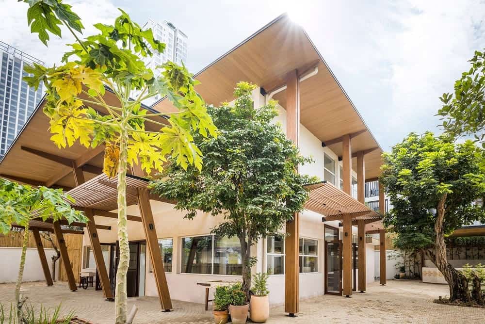 This is an exterior view of the house with bright beige exterior walls and multiple windows. These are then complemented by the large wooden structures and panels attached to it as well as the trees and shrubs of the landscaping.