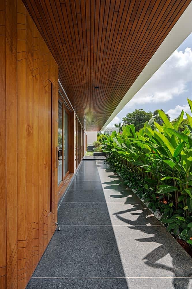 This is a close look at the front of the house with a walkway of gray tiles topped with a wooden ceiling. These are then complemented by the row of tropical plants on the side across from the glass walls.