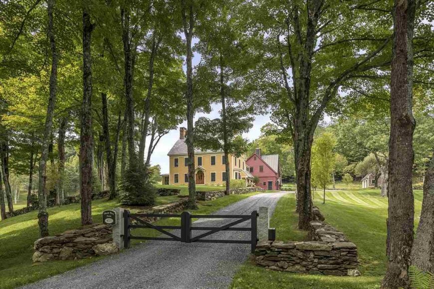 This is outer gate of this Country-style landscaping. This gate is more for aesthetic rather than security with its low stone walls on either side. This opens to a graveled driveway that is shaded by the multiple tall trees around the area dotting the grass lawns.
