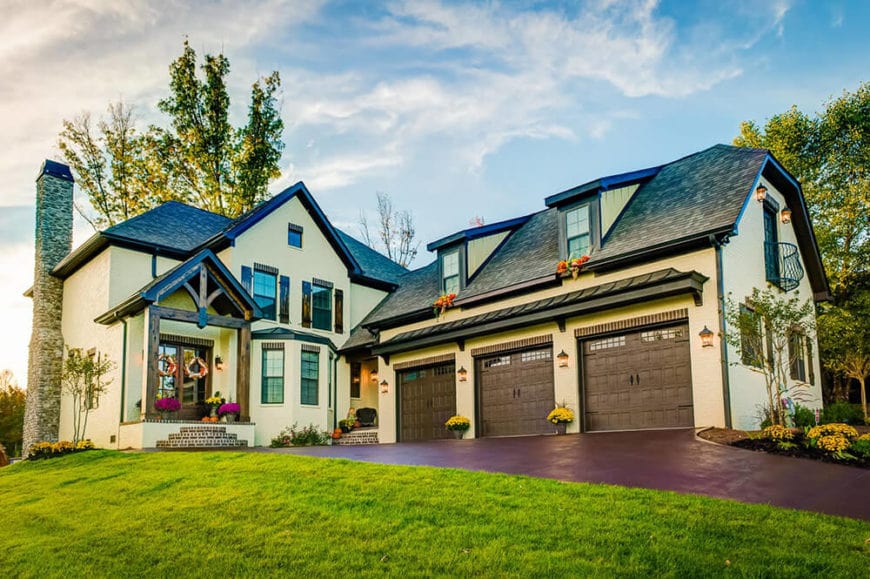 The Country-style landscaping of this home aims to highlight the natural beauty of the home instead of veer attention from it. The earthy driveway and walkway lead to three brown garage doors that are adorned with bright flowers matching those on the main entryway.