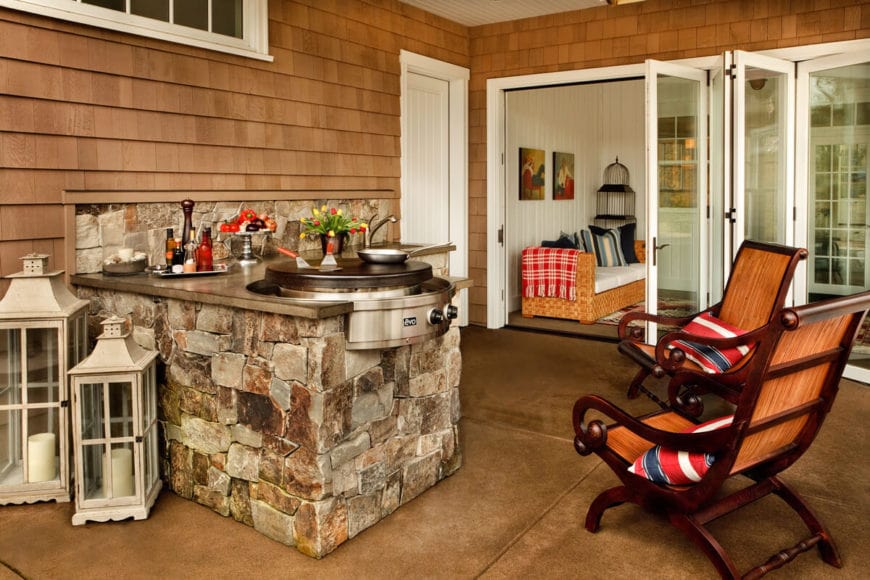 This is an outdoor area right outside of the house complementing it with the charming wooden shingles of the exterior walls. This goes well with the textured stone peninsula and backsplash of the outdoor kitchen. This is paired with a couple of dark wooden chairs that stand out against the gray outdoor tiles of the floor.