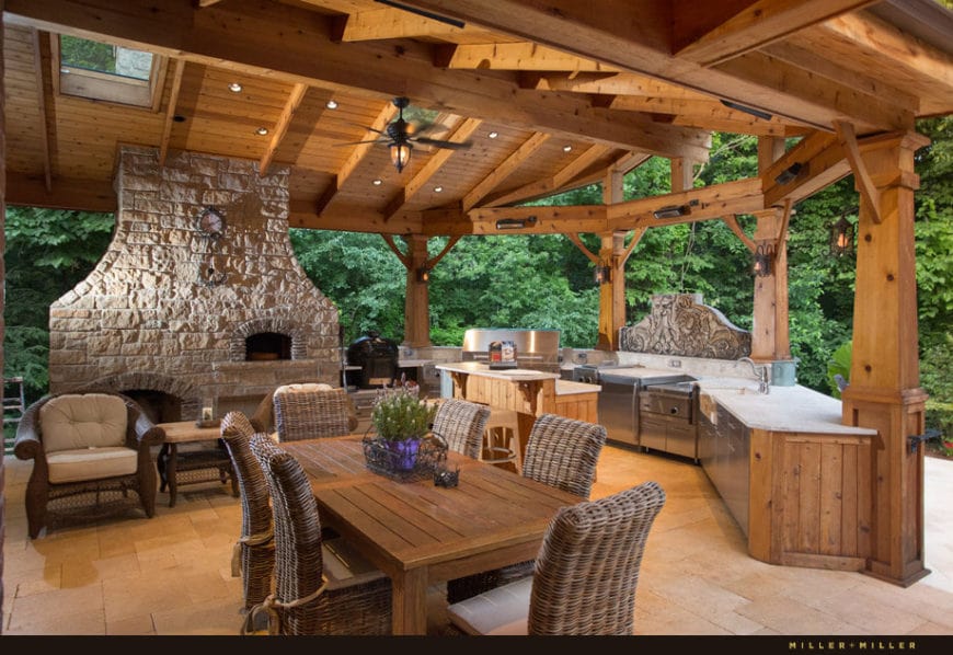 This is the outdoor kitchen and dining area of this home that has a charming wooden cathedral ceiling with exposed wooden beams complemented by the textured stone pillar of the fireplace. The open side of this area features the lush greenery of the Country-style landscaping surrounding the area.