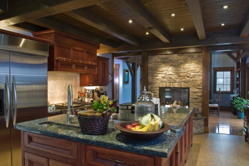 This is a dark kitchen with dark brown wooden cabinetry on its kitchen island and the wooden structure that houses the oven and fridge. These are matched by the dark wooden ceiling with exposed wooden beams and are all complemented by the textured stone fireplace.