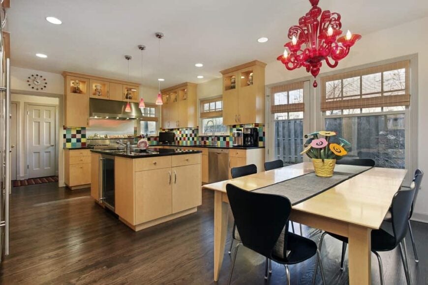 This eclectic dine-in kitchen has a colorful rainbow variety of small backsplash tiles to complement the light wooden cabinetry and black countertop of the L-shape kitchen. This is paired with a small kitchen island of the same tone making it stand out against the dark hardwood flooring.