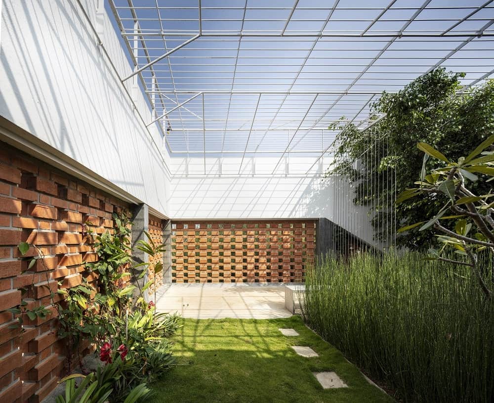 This is a close look at the indoor garden of the house with a glass ceiling supported by metal beams that bring natural lighting to the grass lawn, shrubs and trees complemented by the red brick walls.