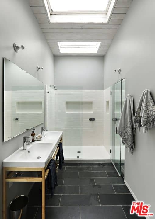 Galley bathroom with black brick flooring and a shiplap ceiling fitted with a pair of skylights. It is filled with a walk-in shower and dual sink washstands under the frameless mirror.