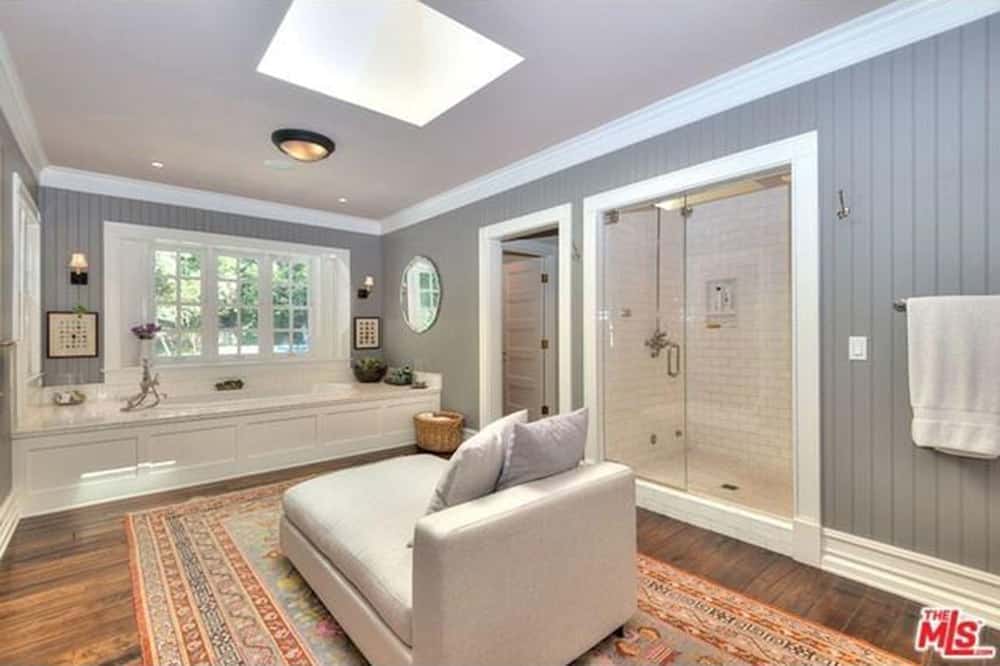 This primary bathroom boasts a walk-in shower and a deep soaking tub facing the cozy chaise lounge on a red printed rug underneath the skylight. It has wide plank flooring and gray beadboard walls mounted with a round mirror along with a pair of sconces and artworks.