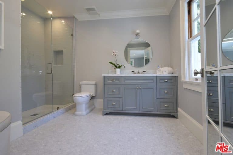 A mirrored door opens to this primary bathroom with a walk-in shower and a traditional toilet that sits on hex tile flooring, It includes a gray vanity paired with a round mirror that's fixed against the gray walls.