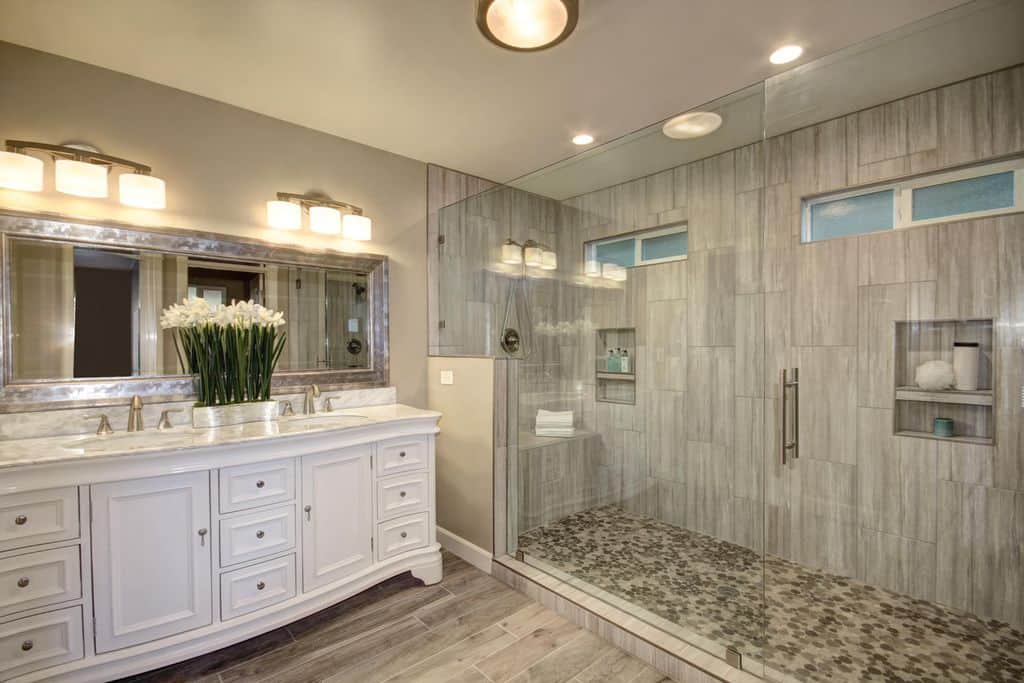 A rectangular mirror is fixed above a marble top vanity that sits next to the walk-in shower with inset shelves and a tiled bench. It is illuminated by glass sconces along with a flush mount light.