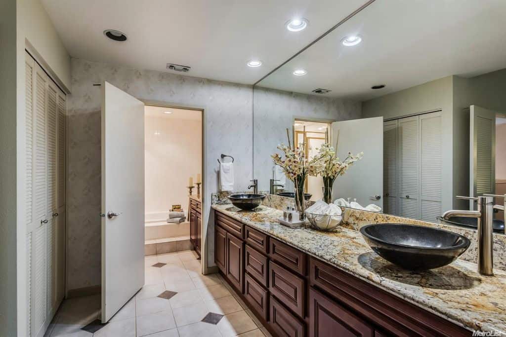 Long and narrow bathroom with a dual vessel sink vanity and a built-in wardrobe fitted with louvered windows. There's a white door in the middle that opens to the deep soaking tub accompanied by another granite top vanity.