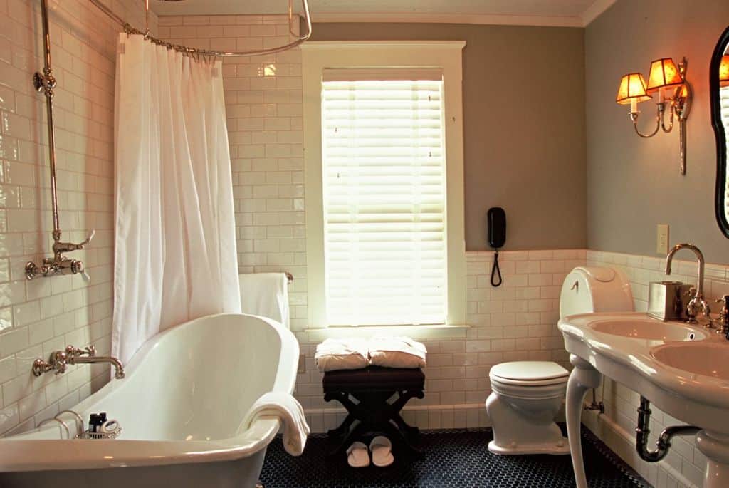 This gray bathroom features a unique toilet and a freestanding tub integrated with a shower. It includes a dark wood stool and a porcelain washstand lighted by warm sconces.