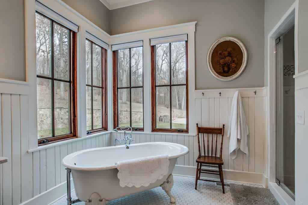 Country style bathroom showcases beadboard walls accented with a round floral artwork along with a wooden framed windows overlooking the enchanting forest. It is filled with a walk-in shower and a clawfoot tub that's paired with a wooden chair over hex tile flooring.