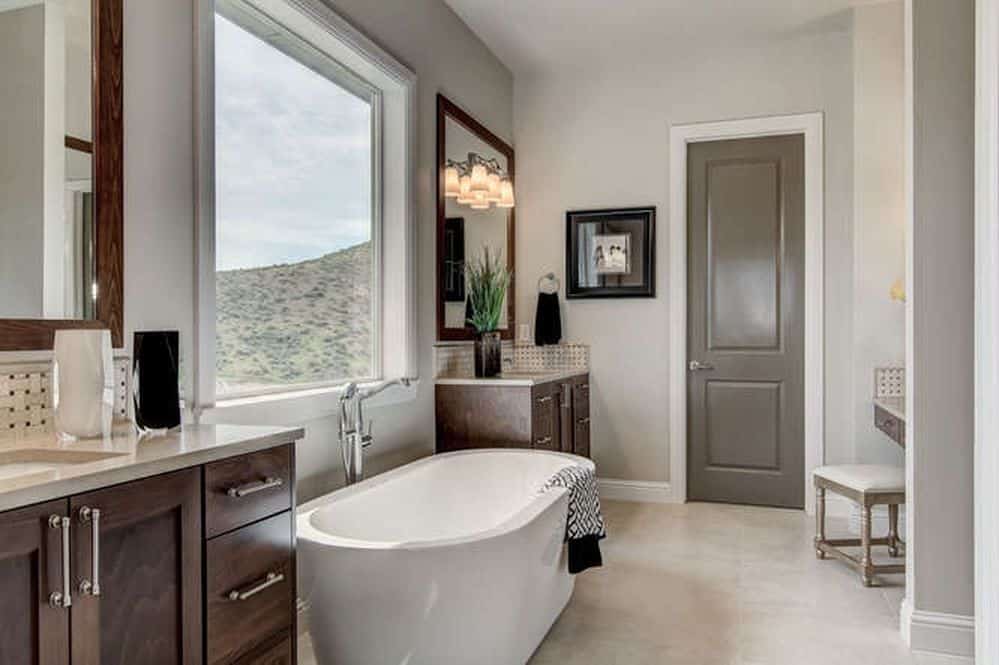 Dark wood vanities flanked a freestanding tub by the picture window with a stunning mountain view. It is accompanied by a black framed wall art and a cushioned stool over beige tiled flooring.