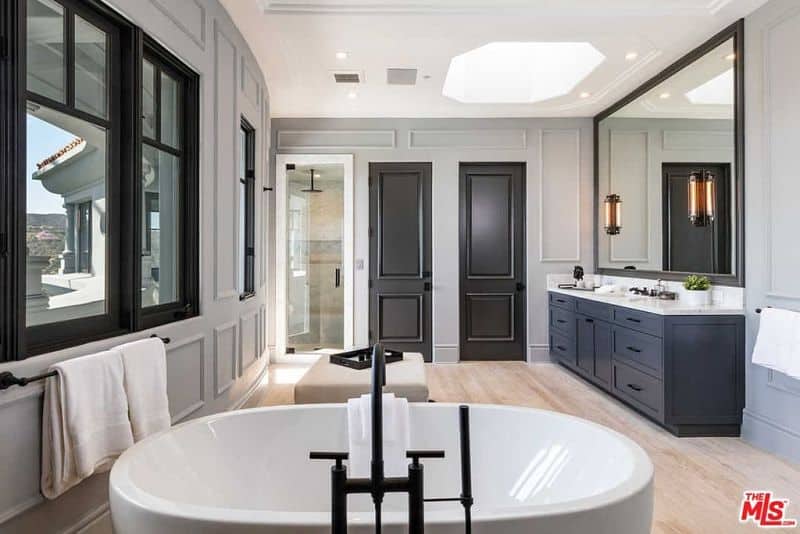 Linear sconces mounted on the large mirror illuminate the marble top vanity that's fixed against the gray wainscoting. It is accompanied by a walk-in shower and a beige ottoman along with a freestanding tub fitted with wrought iron fixtures.