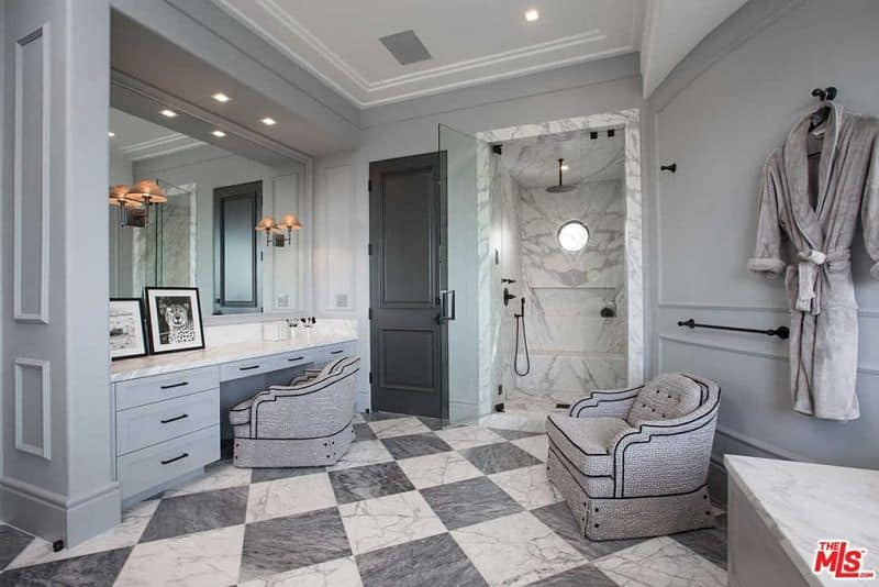 The opposite side of the deluxe primary bathroom showcasing a marble top vanity and charming gray seats along with a walk-in shower that's fitted with an inset shelf and wrought iron fixtures.