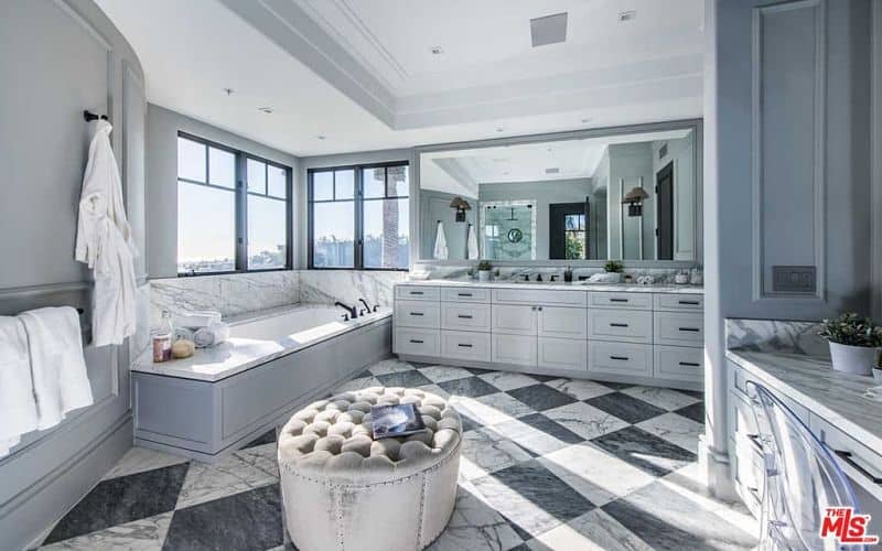 Deluxe primary bathroom with gray vanities and a deep soaking tub by the aluminum framed windows inviting an ample amount of natural light. There's a tufted ottoman in the middle that sits on a striking marble checkered flooring.