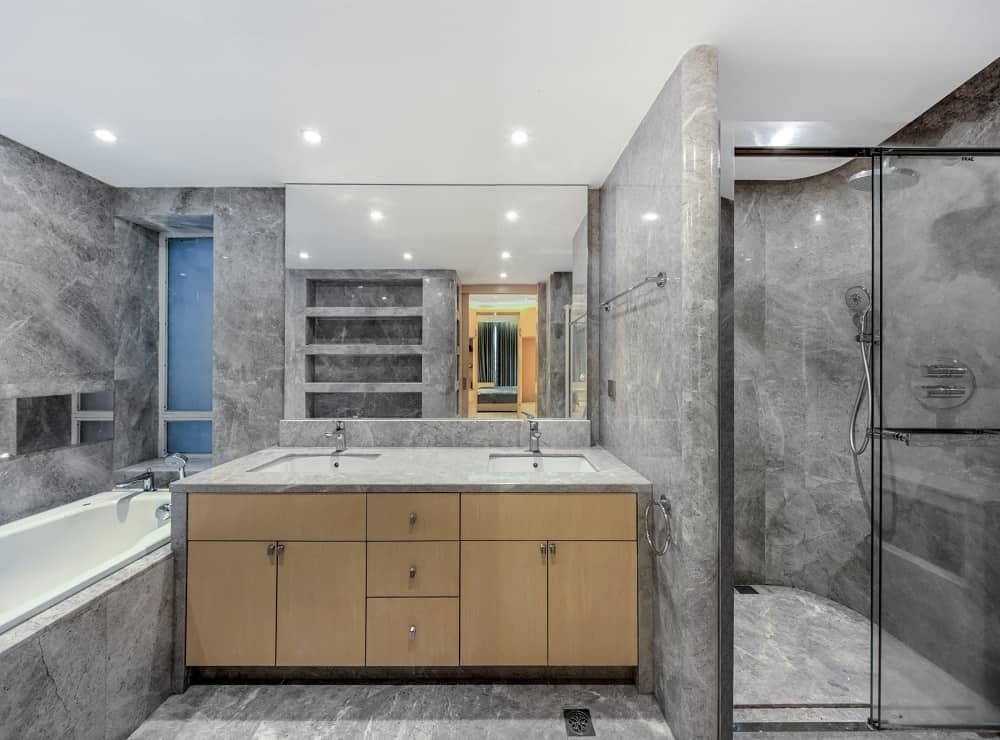 This bathroom has consistent gray marble tones on its floor, walls and the walls of the walk-in shower with a glass door by the wooden vanity.