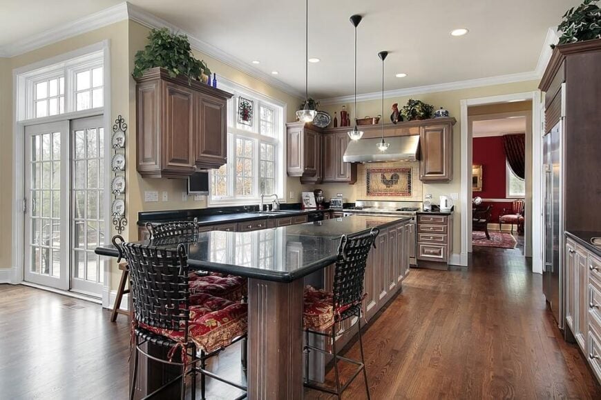The extensive dark wood of this kitchen extends from the hardwood flooring to the wooden shaker cabinets of the beige walls and the large wooden structure housing the stainless steel fridge. It can also be seen on the large kitchen island paired with wrought iron stools to match the black countertop.