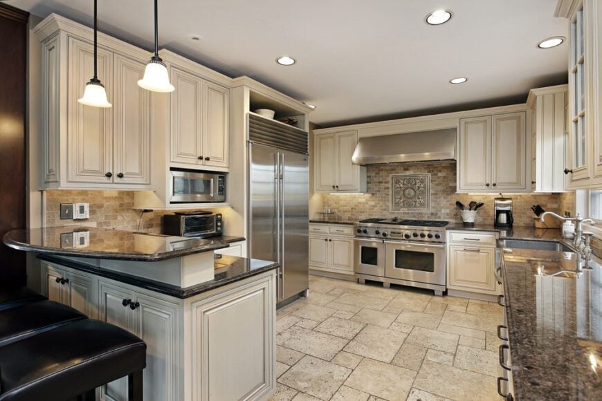 This charming spacious kitchen has an L-shaped peninsula with white white shaker cabinets paired with black countertops and a second tier to serve as a breakfast bar for the black cushioned stools. This is topped with a couple of small pendant lights hanging from the white ceiling.