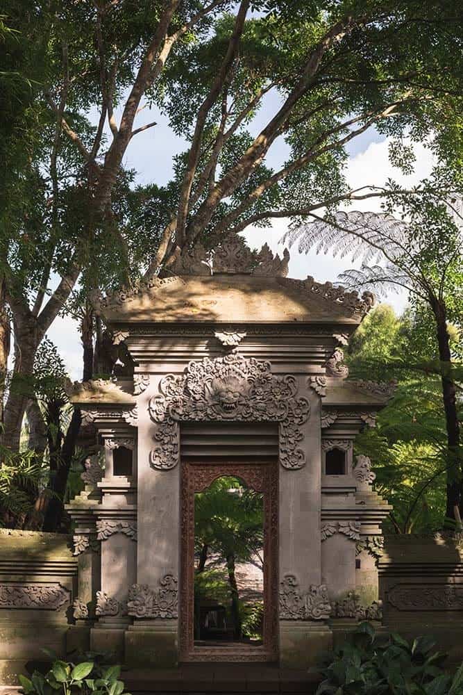 This is a full view of the house's large concrete archway and outer gate with intricate carvings and images on it that gives it a distinct Asian look surrounded by tall trees and shrubs.