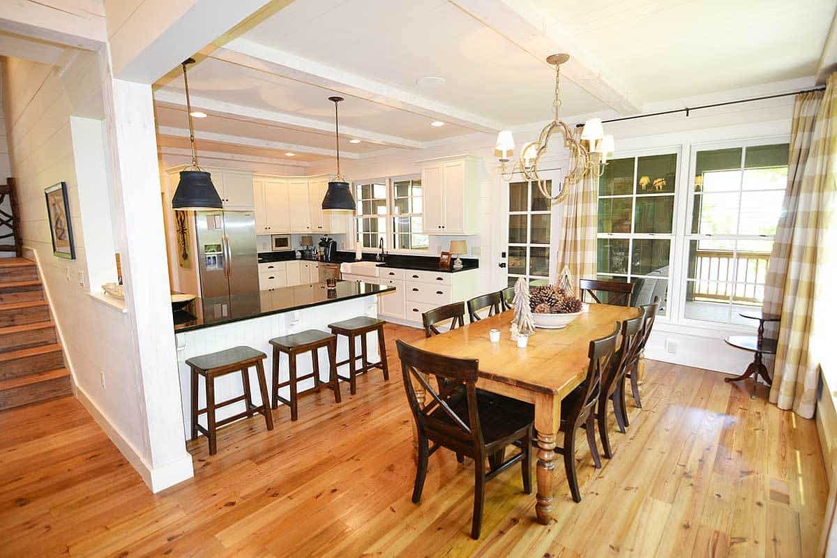 This is an informal dining area beside the kitchen that has a large wooden dining table to match the hardwood flooring. The wooden chairs surrounding it are of a slightly darker hue to contrast the white walls and ceiling that hangs a simple chandelier over the table.