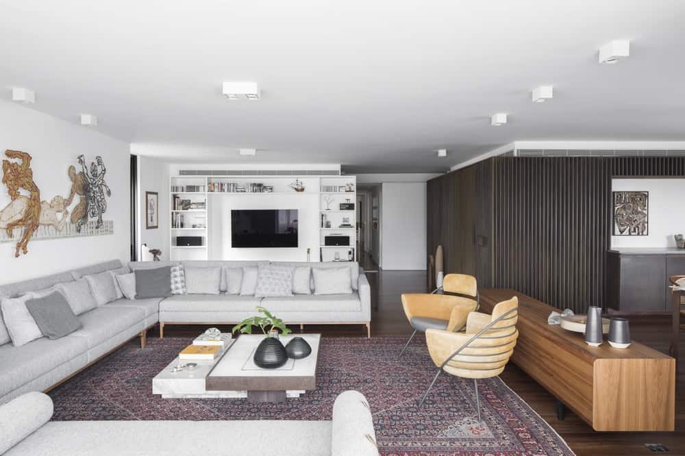 This is a full view of the living room with a large sectional gray sofa on one side with a coffee table in the middle of a large patterned area rug. On the far side is the white cabinetry that houses the TV.