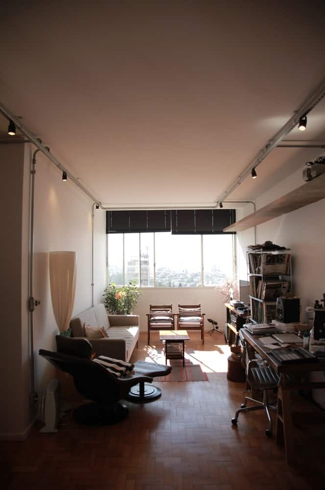 This is the living room that has a sofa on one side and home office on the other side with structures and desk brightened by the modern lighting above and the windows on the far side.