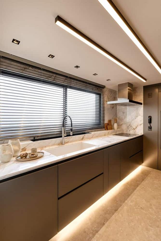 This is a close look at the kitchen with a set of modern dark cabinetry on the wall beneath the window topped iwth a bright counter that houses the faucet area and the cooking area on the far corner. These are then complemented by the modern lighting underneath.