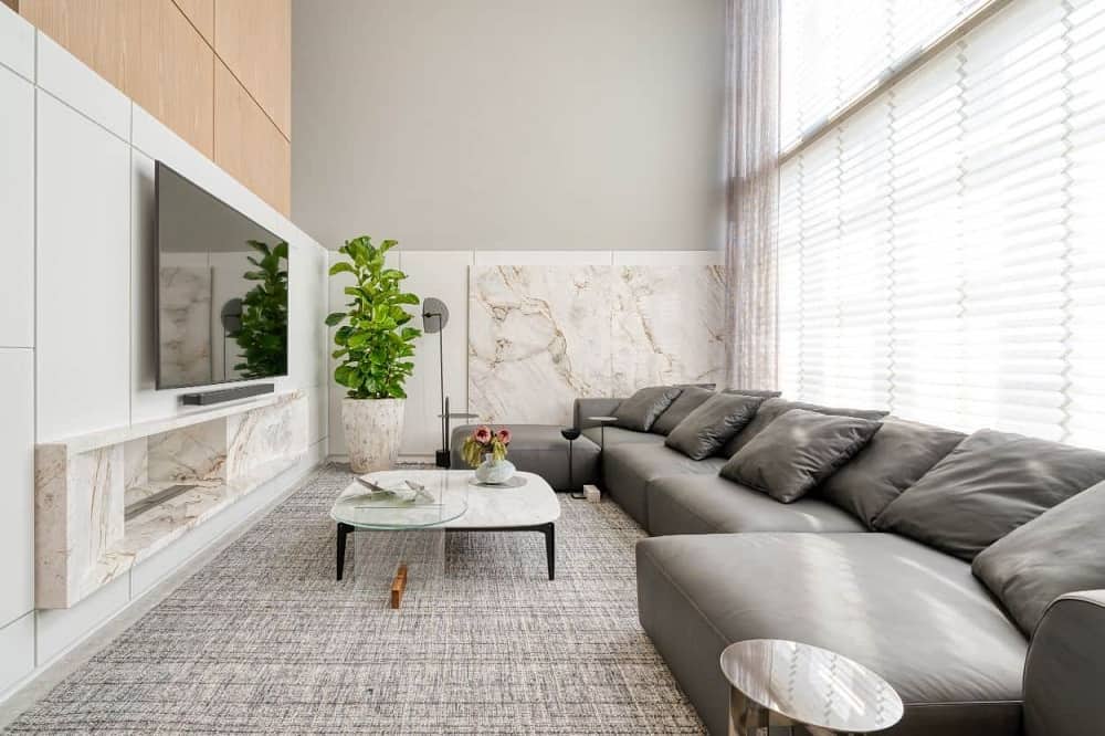 This is a full view of the apartment's living room with a large gray leather sectional sofa across from the wall-mounted TV, modern fireplace and potted plant. These are all complemented by the large glass wall behind the sofa.