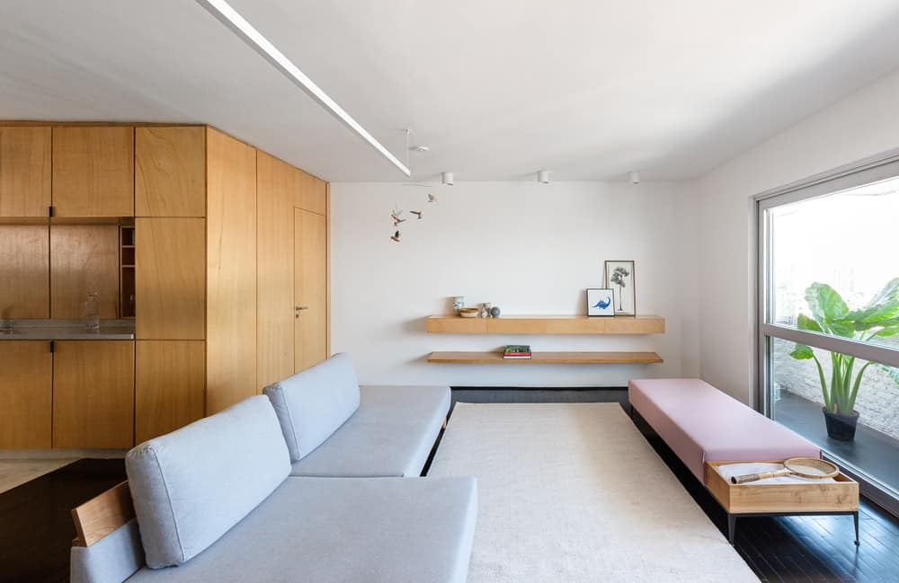 This is a full view of the simple living room that is dominated by a large sectional light gray sofa. There is a set of floating wooden shelves on the far side of the area.