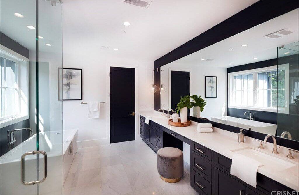 Black and white primary bathroom with a freestanding tub and a dual sink vanity paired with a round velvet ottoman. It is illuminated by recessed ceiling lights along with natural light flowing in from the white-framed windows.