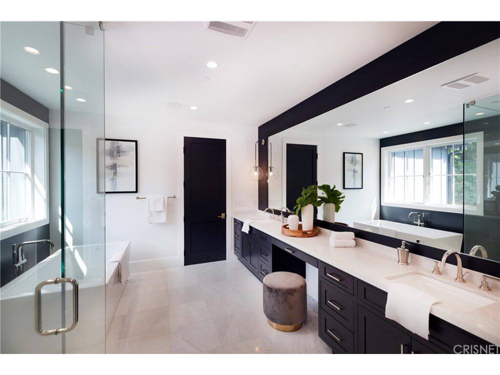 Black and white primary bathroom with a freestanding tub and a dual sink vanity paired with a round velvet ottoman. It is illuminated by recessed ceiling lights along with natural light flowing in from the white-framed windows.