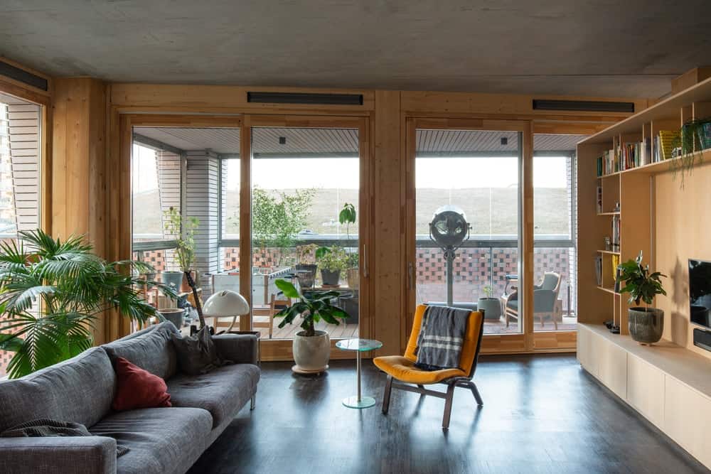 This is a full view of the living room area that has a gray sofa across from a large wooden structure that has shelves and cabinets. These stand out against the dark hardwood flooring adorned with potted plants.