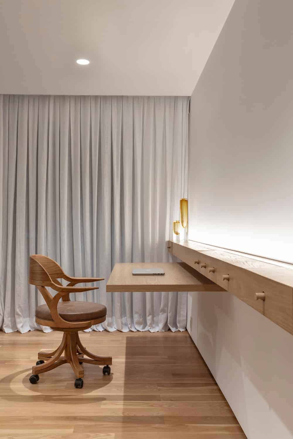 A cozy home office with a round back swivel chair and a built-in desk that matches the hardwood flooring. White drapes along with ambient lighting create warmth in the room.