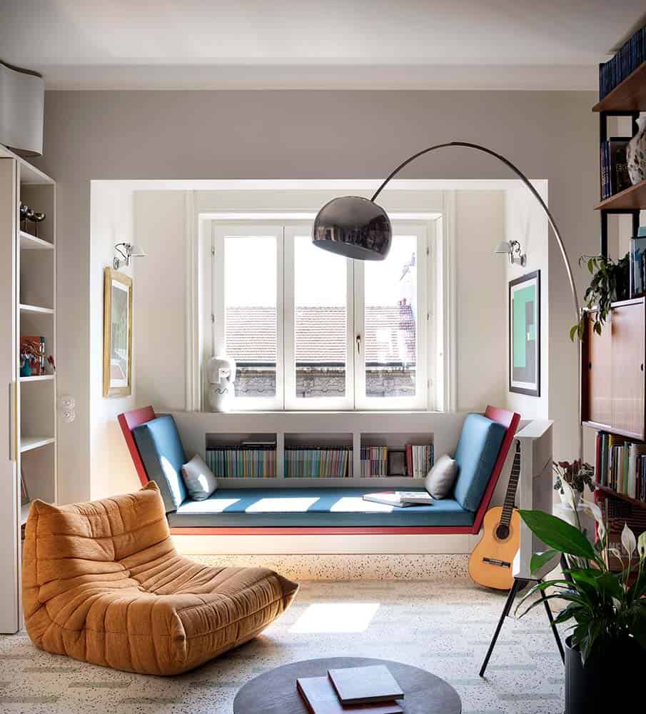 This is a close look at the living room with multiple built-in book shelves on the walls and the space underneath the windows that is also fitted with a cushioned built-in bench as a reading nook brightened by the natural lights of the windows.