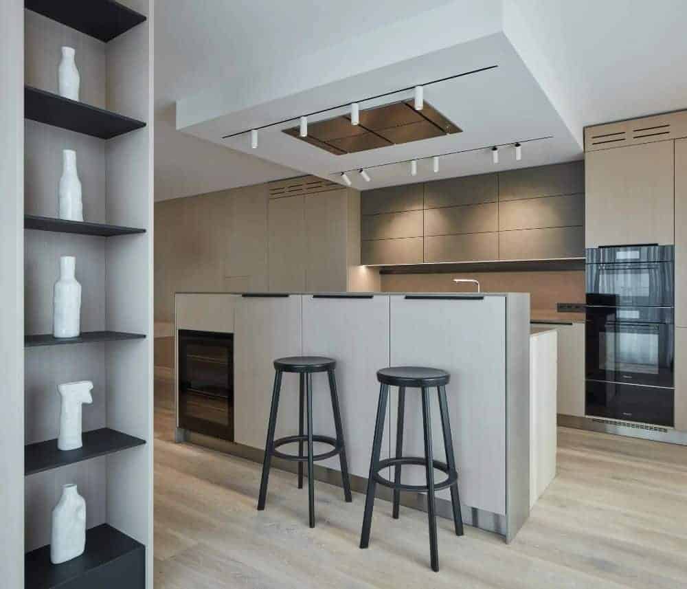 This is a look at the kitchen with a breakfast bar to its kitchen island with a second tier level. The cooking area is topped iwth a modern vent hood with modern lighting on the sides. On the far wall, the black appliances stand out.