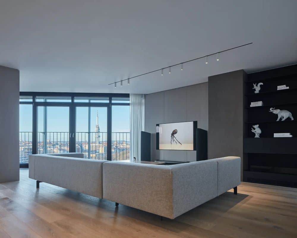 This is an interior view of the apartment's minimalist living room with a large sectional sofa across from the TV. These are then complemented by the glass windows that have panoramic views outside with a balcony beyond the glass doors.