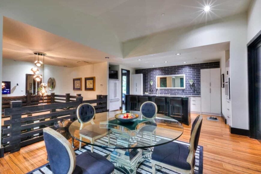 The dining area offers a round glass top table and blue tufted chairs matching with the striped rug.