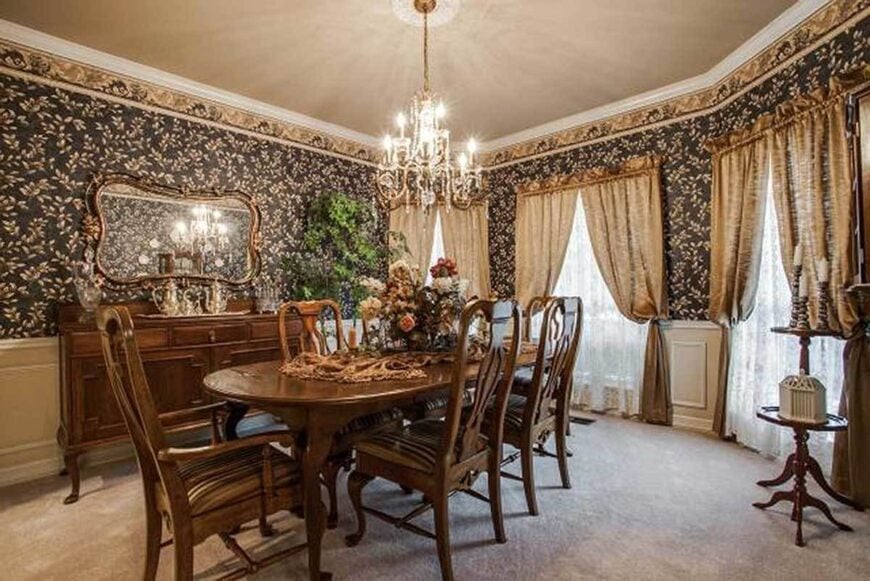 The formal dining room has a classic look with its classy wallpaper, beige wainscoting, and wooden furniture sitting over the carpet flooring.