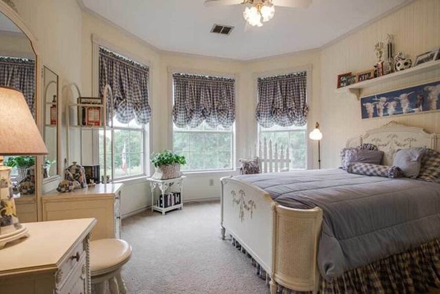 This bedroom has a classy white vanity and a comfy bed with a blue checkered skirt matching with the bay window's roman shades.