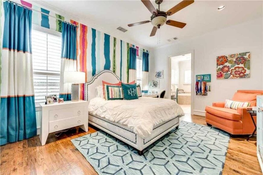 This bedroom has rich hardwood flooring and light beige walls adorned by colorful striped wallpaper and floral painting.